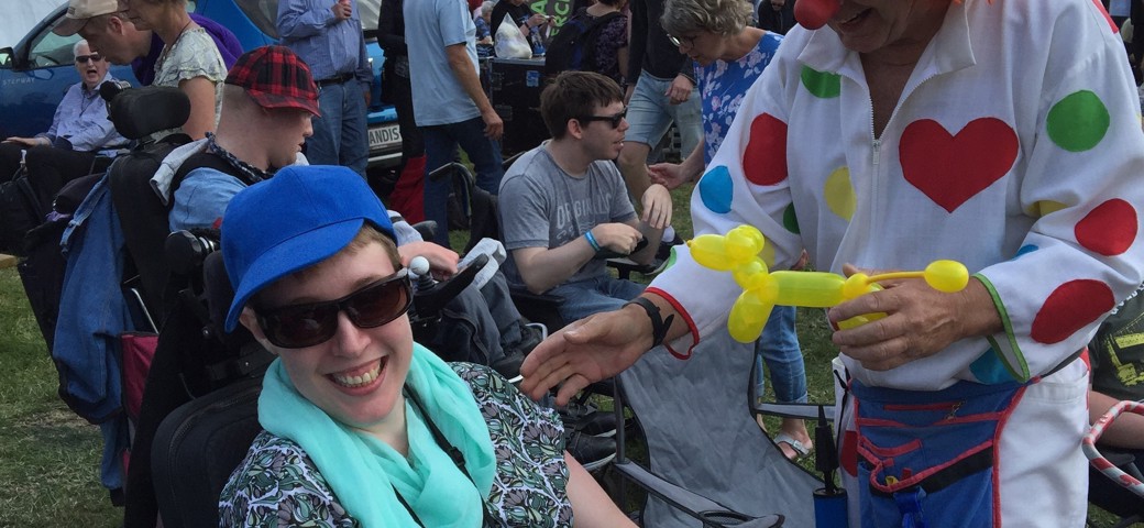 Kvinde sidder med solbriller og smiler, mens en mand klædt ud som en klovn er ved at lave et ballondyr